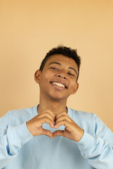 Vertical image of a cheerful brown skinned teenager wearing a pastel blue sweatshirt making a heart gesture with his hand on a light brown background with copy space, feeling of love with his hands