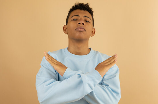 Brown skinned Latino teenager wearing a pastel blue sweatshirt, with an angry face with his hands crossed making a rejection expression on a light brown background with copyspace
