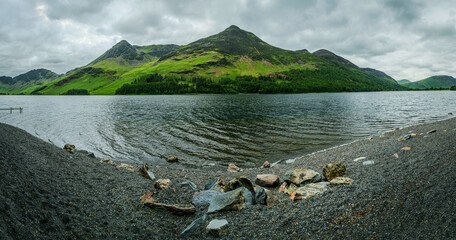 Panoramic view from the shore of Buttermere Lake shows rocks scattered along the pebble beach,...