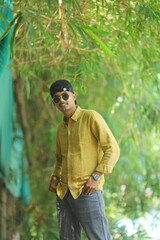 A stylish young man wearing a mustard yellow shirt, grey jeans, and sunglasses poses confidently in...