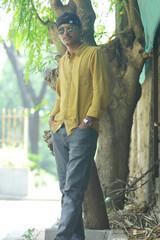 A stylish young man wearing a mustard yellow shirt, grey jeans, and sunglasses poses confidently in...