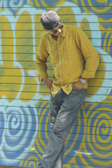 A stylish young man wearing a mustard yellow shirt, grey jeans, and sunglasses poses confidently in...