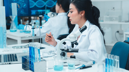 Asian female scientist in lab coat working on a laptop and taking notes in a modern laboratory....