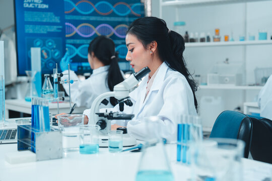 Asian female scientist in lab coat working on a laptop and taking notes in a modern laboratory. Scientific research environment with test tubes, beakers, and microscope in the background.
