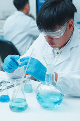 Fototapeta premium Asian male scientist wearing safety goggles and gloves working with blue liquid in a laboratory. Other researchers are working in the background in a clean, modern scientific environment.