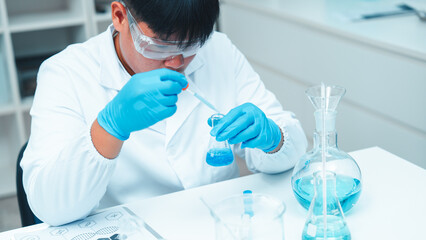 Asian male scientist wearing lab coat and safety goggles using pipette to transfer blue liquid in...