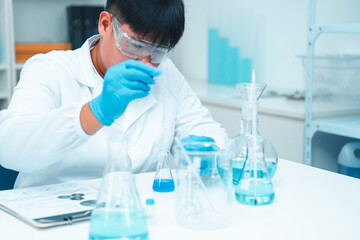 Asian male scientist wearing lab coat and safety goggles using pipette to transfer blue liquid in...