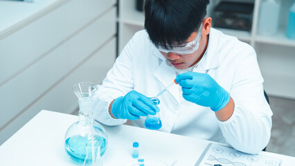 Asian male scientist wearing lab coat and safety goggles using pipette to transfer blue liquid in...