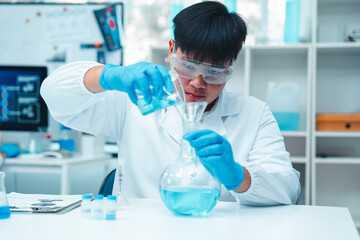 Male scientist in white lab coat and protective eyewear carefully pouring blue liquid into a glass...