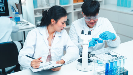 Two scientists in white lab coats smiling and collaborating in a laboratory. One holds a test tube while the other writes notes beside a microscope. Teamwork and positive energy in scientific research
