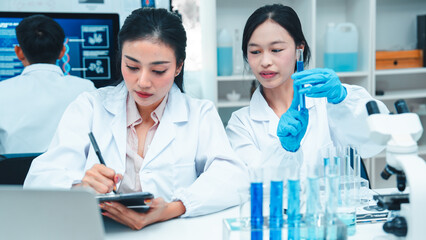 Two female scientists in lab coats working together in a science laboratory. One takes notes on a...