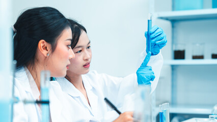 Two female scientists in lab coats working together in a science laboratory. One takes notes on a...