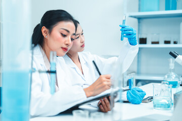 Two female scientists in lab coats working together in a science laboratory. One takes notes on a...