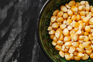 Yellow corn kernels in bowl. Closeup of popcorn maize on black marble table. Raw food grains background. Organic dry corn seeds. Healthy snack preparation. Vegetarian ingredient ready to cook.
