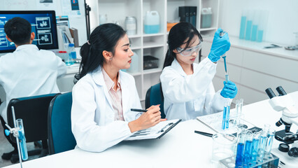 Two female scientists analyzing test tube samples in a laboratory setting. One supervises while the other conducts an experiment. Concept of science education, teamwork, and research collaboration.
