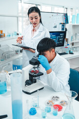 Male scientist examining a specimen under a microscope while a female scientist records...