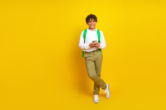 Cheerful schoolboy using smartphone while standing with yellow background, showcasing back-to-school essentials in style