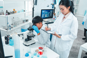 Male scientist examining a specimen under a microscope while a female scientist records...
