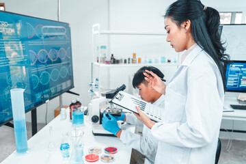 Scientist observes a sample under a microscope while a female colleague records data on a clipboard...