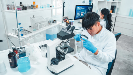 Young male scientist in lab coat examining a petri dish with a magnifying glass in a laboratory....