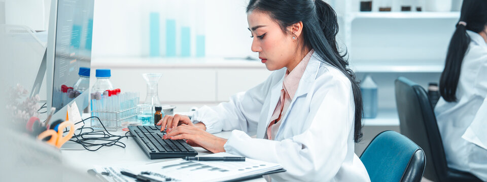Female scientist in lab coat typing on a keyboard while analyzing research data on a computer. Laboratory equipment and chemical samples are visible, showing a scientific and focused work environment.