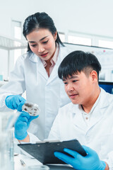 Female senior scientist supervising young male researcher in a modern lab. She reviews data on a clipboard while he listens attentively. Professional teamwork in scientific research setting.