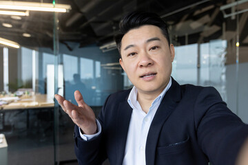 Close-up photo of a young Asian male businessman in the office talking to a camera he is holding in...