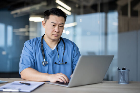 Serious and focused young Asian male doctor sitting at his desk in a blue uniform and working on a laptop