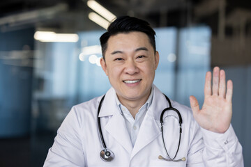 Close-up photo of a smiling young Asian doctor in a white coat and with a stethoscope in a clinic,...
