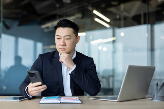 Asian young male businessman sitting in the office at his desk, looking thoughtfully at his phone