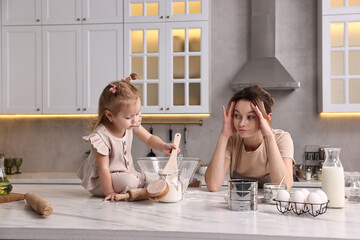 Tired housewife cooking with her little daughter at marble table in kitchen