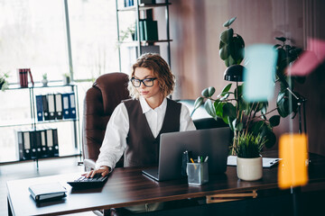 Focused professional businesswoman in a modern office working on a laptop while calculating data...