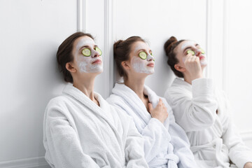 Obraz premium Happy women with facial masks and cucumber slices in bathrobes after spa procedures near white wall