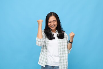 Fototapeta premium Young asian woman raising arms with clenched fists celebrating success, having cheerful mood feeling happy, closing eyes, wearing casual clothes, standing isolated on blue background.