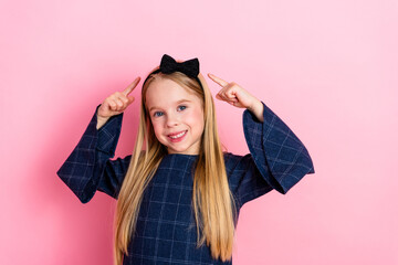 Smiling girl with long blonde hair pointing to her bow, wearing a blue casual dress, posing against...