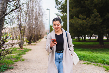 Young woman influencer creating content for social media walking in a park listening to music with headphones