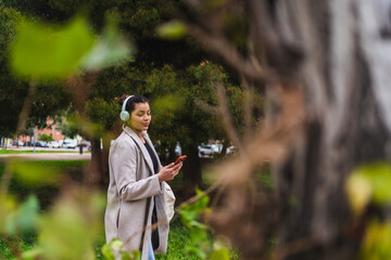Young stylish influencer woman walking in a park listening music with headphones and using smartphone