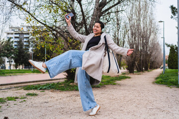 Young stylish influencer dancing and listening to music with headphones and smartphone in a park