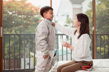 A boy in fencing gear with their mother preparing food and sports equipment