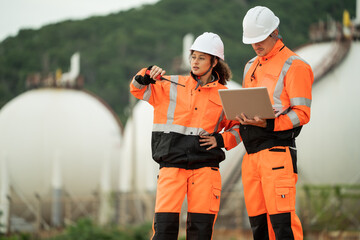 Two refinery workers discuss data on laptop, Backgroud of large storage tank, engineer communicate using walkie-talkie, industrial site operation