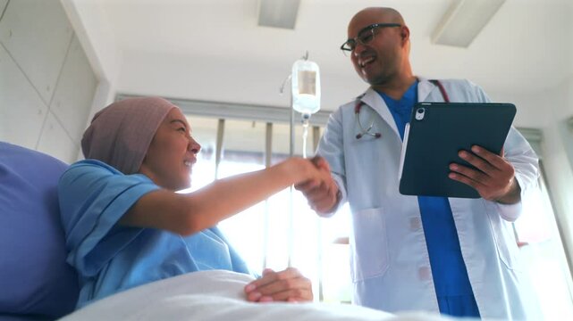 Asian woman patient describes her symptoms to an oncologist while sitting on a bed and looking at a man doctor in a lab coat in a hospital ward, health and medical care concept