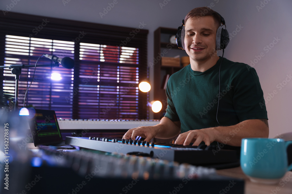 Poster smiling man in headphones working with midi keyboard at table in home studio, low angle view