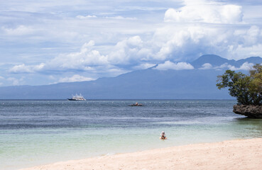 Solitary woman bathing in turquoise waters on a quiet tropical beach with a view of distant mountains, dramatic clouds and sailing boats on the horizon.