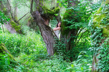 woman in white dress blends into vibrant green forest, standing between ancient mossy trees –...