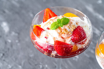Ice cream with whipped cream, strawberries and peach in a glass tall glass on a gray background....