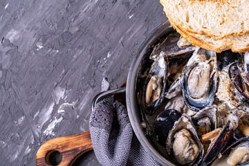 Mussels in shells cooked in a creamy sauce in a saucepan with toasted toast on a gray background....
