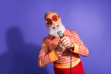 Charismatic senior man in retro style attire performs singing gesture against a vibrant purple...