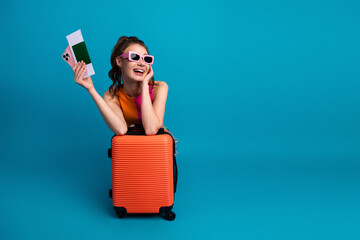 Cheerful young woman with summer style sitting on an orange suitcase holding a smartphone and...