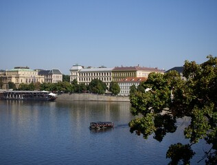 Naklejka premium Serene waters of a European city reflecting historic architecture under a clear sky, with a boat cruising by.