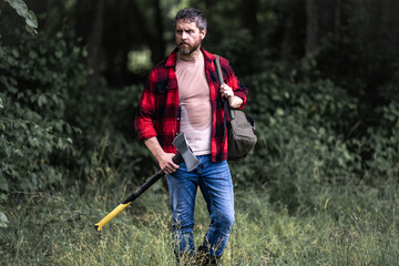 Confident lumberjack works in summer forest. Man with an ax. Woodcutter in plaid shirt in forest....